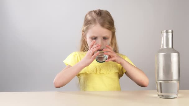 Klein meisje in een gele jurk op een witte achtergrond drinkt water uit een glas. — Stockvideo