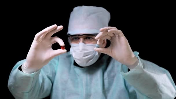 Doktor v chirurgických šatech v lékařské masce s brýlemi ukazuje na dvě tablety červené a modré. Doktor drží dva prášky v bílých lékařských rukavicích. na černém pozadí. — Stock video