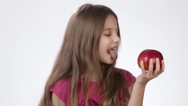 Klein meisje op wit eet een grote rode appel. Een meisje bijt in een stuk rode as en proeft het.. — Stockvideo