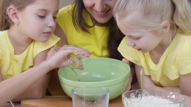 Mama z dziećmi gotuje w kuchni. Dziewczyna wlewa olej do ciasta w zielonej misce. Mama z dziećmi w kuchni w żółtych ubraniach — Wideo stockowe