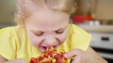 Küçük kız bir dilim pizzayı yakından ısırıyor ve lezzetli bir şekilde pizza yiyor. Kız iştah açıcı bir şekilde pizza çiğniyor..