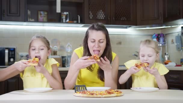 Mamá con dos niñas comiendo pizza en casa en la cocina. Los niños y mamá comen deliciosa pizza en ropa amarilla . — Vídeos de Stock