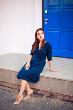 Koyu mavi elbiseli, kırmızı saçlı bir model mavi kapıda poz veriyor.
