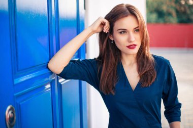 Koyu mavi elbiseli, kırmızı saçlı bir model mavi kapıda poz veriyor.