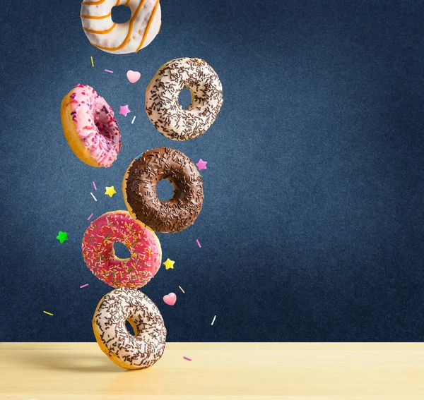 Fliegende Donuts. Mischung aus bunten süßen Donuts mit Streusel fällt auf den Tisch auf dunkelblau strukturierten Hintergrund. — Stockfoto