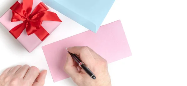 Caja de regalo rosa con lazo rojo y manos masculinas escribiendo carta de amor, tarjeta de felicitación para el día de San Valentín o invitación a la boda. Vista superior. Fondo blanco . —  Fotos de Stock
