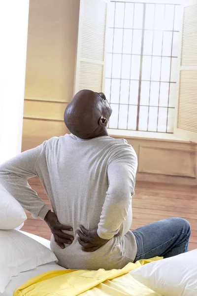 Афроамериканец с сильной болью в спине сидит на кровати в своей роскошной спальне — стоковое фото