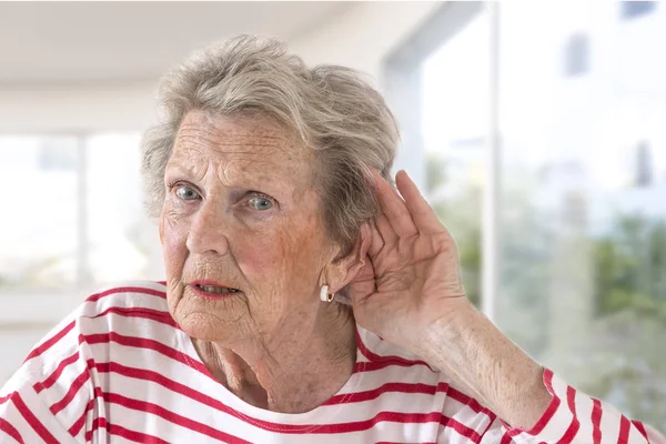 Señora anciana con problemas auditivos debido al envejecimiento sosteniendo su mano al oído mientras lucha por escuchar, vista del perfil en el fondo de grandes ventanas — Foto de Stock