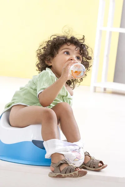Kleine baby, zit op een blauwe pot en drinken water uit een fles binnenshuis achtergrond — Stockfoto