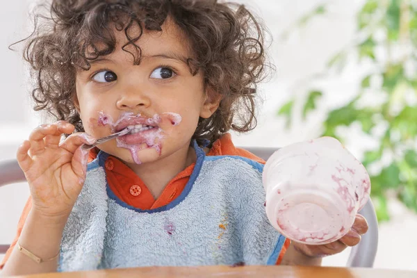 Criança sentada em cadeira alta e comendo iogurte grego. Bebê aprendendo a comer e tem iogurte no rosto e cabelo — Fotografia de Stock