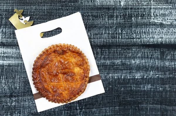 Epifanía Duodécima Noche pastel francés galette des rois hecha de hojaldre, con el encanto en el interior, en la corona bolsa de hornear apoyado dentro de fondo de madera negro — Foto de Stock