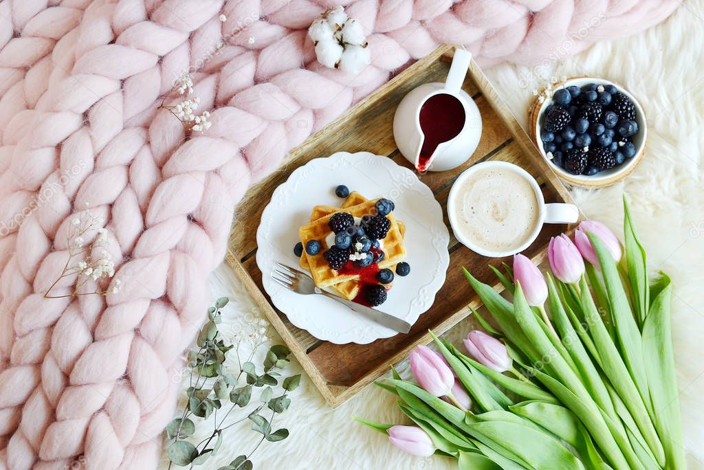 Cup with cappuccino and homemade Belgian waffles with strawberry sauce and berries, pink pastel giant blanket, fur, bedroom, pink flowers tulips, spring, woman day morning concept 