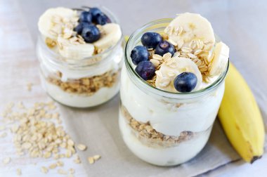 Vitamini sağlıklı kahvaltı Yoğurtlu yulaf gevreği, muz ve yaban mersini Beyaz ahşap arka plan püresi Cam Kavanoz yararlı gıda