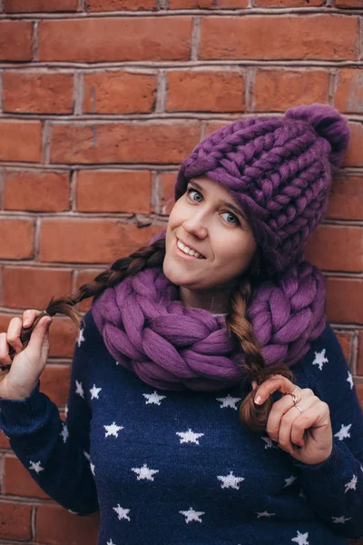 Brunette woman in purple knitted hat, knitted infinity scarf and blue sweater with white stars on it make posing with brick wall on the background