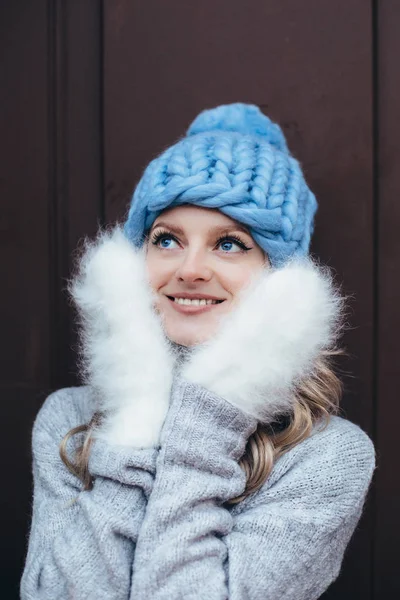 Young blonde woman in blue knitted hat, white fluffy mittens and grey sweater make posing with wooden door on the background