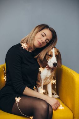 Arka plan gri duvar ile sarı sandalyeye beagle köpek yavrusu siyah elbiseli genç kadın hugs