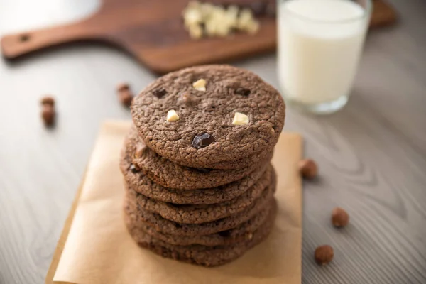 Čokoládové sušenky s lískovými oříšky, bílé čokolády a tmavá čokoláda na pergamen a sklenici mléka, dřevěné pozadí — Stock fotografie