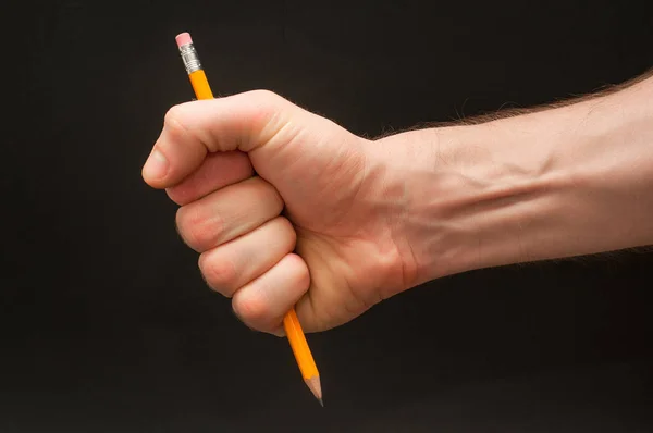 Braço masculino direito segurando lápis de cor laranja simples em posição ameaçadora em um fundo preto largo abstrato isolado . — Fotografia de Stock