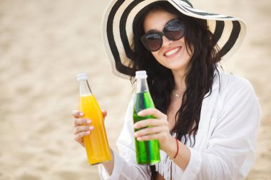 Kumsalda oturuyor ve sarı ve yeşil kokteyl kumsalda tutan genç kadın