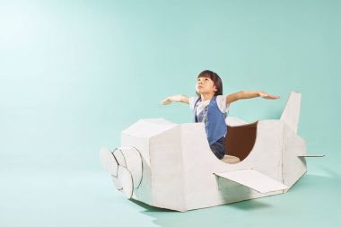 Little cute girl playing with a cardboard airplane. White retro style cardboard airplane on mint green background . Childhood dream imagination concept . clipart