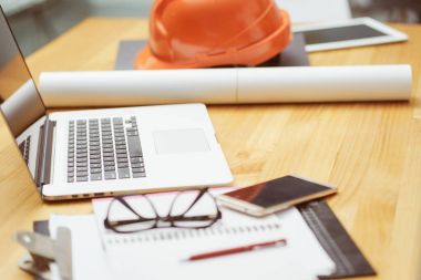 Dizüstü, gözlük, not defteri, telefon, blueprint ve inşaat sahasında masada mühendis, foreman ve işçi, seçici için yığınlama turuncu güvenlik kask odaklanarak .