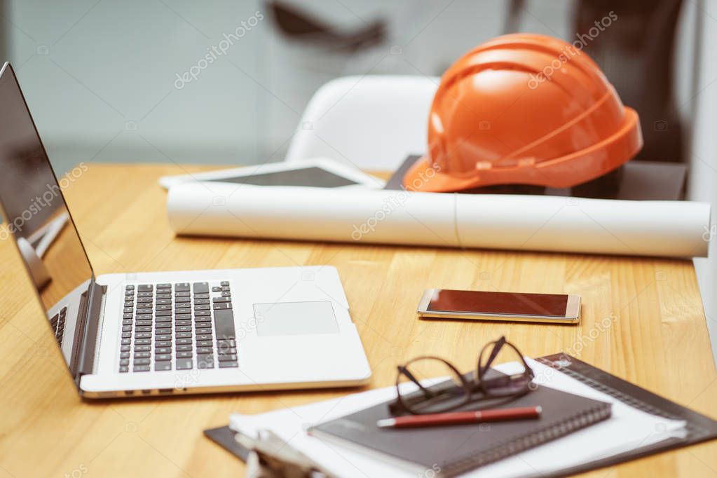 Laptop ,glasses, note book ,phone ,blueprint and orange safety helmet stacking on table at construction site for Engineer, foreman and worker , selective focusing .