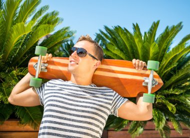 Yeşil palms karşı yaslanmış ve onun longboard - güneş gözlüğü takıyor ve güneşli gün zevk genç adam - spor açık ve yaz konsepti holding yakışıklı adam
