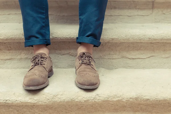 Young man wearing brown chamois shoes with suite and blue pants on stones background in a cloudy day. Sitting hipster guy with old elegant shoes and typical turn-up jeans. Main focus on laces