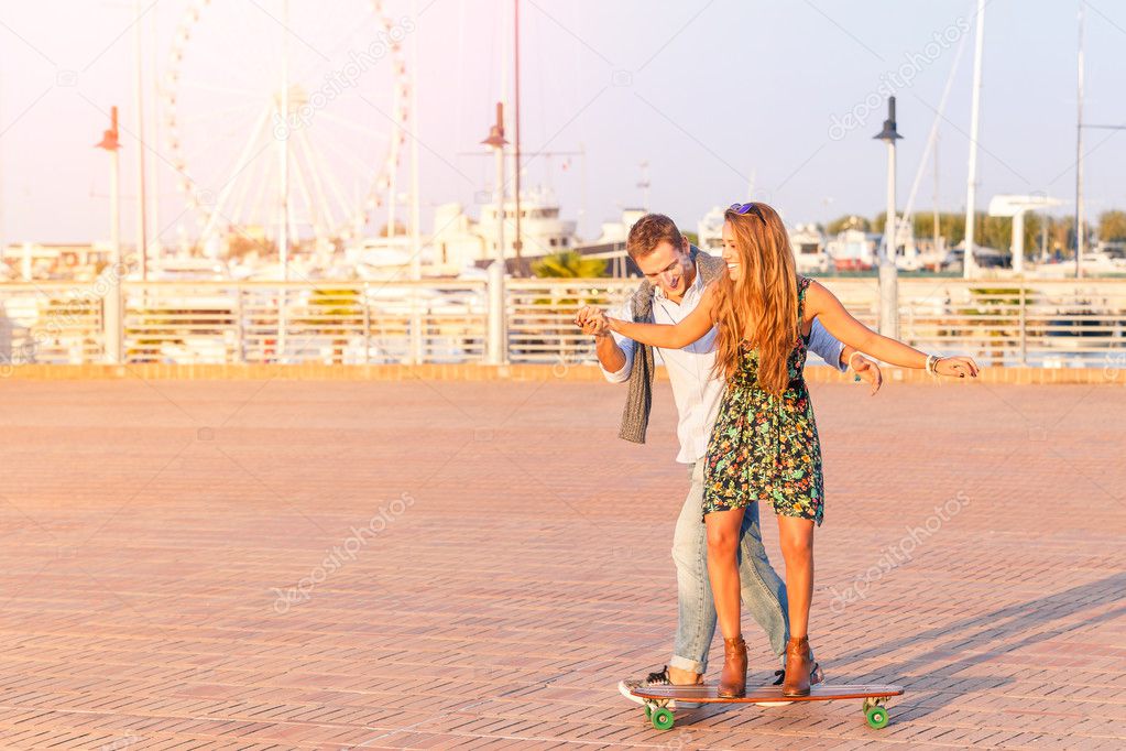 Young hipster best friends having fun at harbor during a sunny day with ferris wheel and boats background. Guy teaching going by longboard to his girl. Lifestyle concept enjoying happy moment together