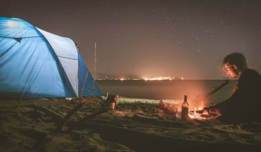 Genç bir adam sahilde çadır izle kamp ateşi ile kırmızı şarap cam önünde oturuyor. Yaz macera güzel doğada en iyi arkadaşlarından. Çadır ana odak ile uzun pozlama fotoğraf.