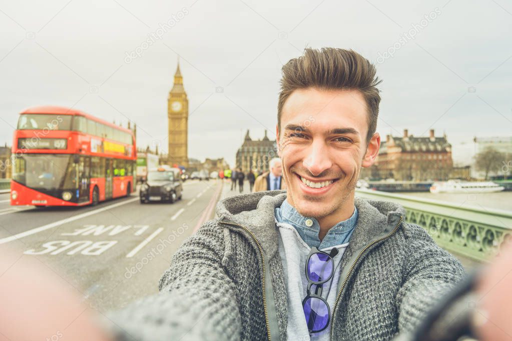 Smiling guy take selfie photo during travel in London, England. Traveler man in front of Big Ben Tower taking memory pic with iconic england red bus. Happy people concept wandering around the world.