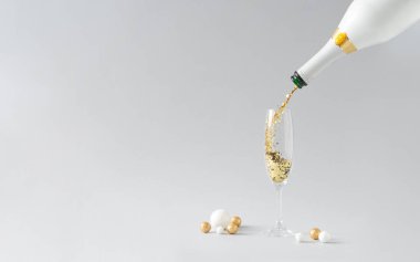 Champagne bottle pouring golden glitter into glass with white and golden decoration against grey background. Celebration minimal Christmas party.