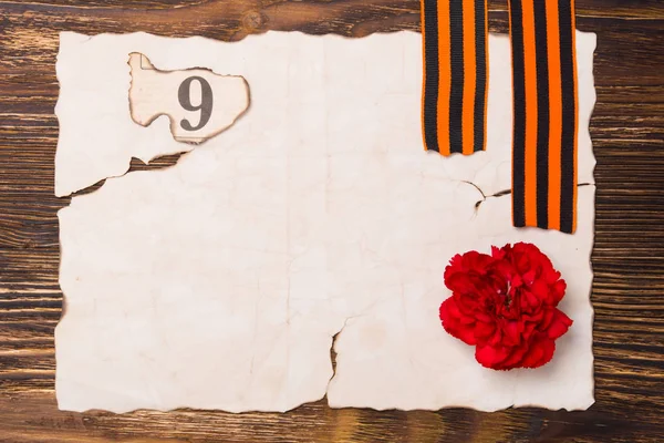 A burnt sheet of paper on the table, with a red carnation and a number 9, and two St. George ribbons — Stock Photo, Image