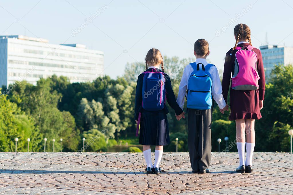 Three pupils in school uniform stand on the street with backpacks, rear view
