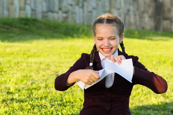 Flicka i skoluniform sitter på gräset och sliter en bit papper — Stockfoto