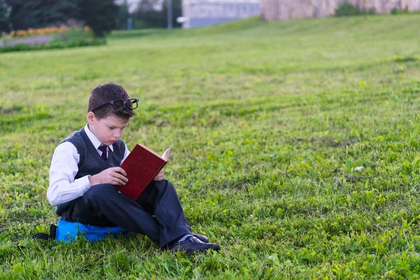 Le garçon en uniforme scolaire est assis sur un sac à dos et lit un livre sur un fond d'herbe verte — Photo