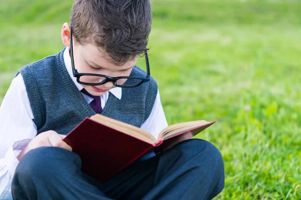 Schüler der Schule, sitzt mit Brille auf dem Rasen und liest ohne Ablenkung ein Buch — Stockfoto