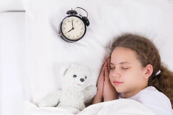 Kız saatin yanında battaniye altında beyaz bir yatakta uyuyor — Stok fotoğraf