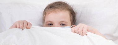 Küçük bir kız tıbbi maskeli, yatakta kendini enfeksiyondan korumak için sakladığını