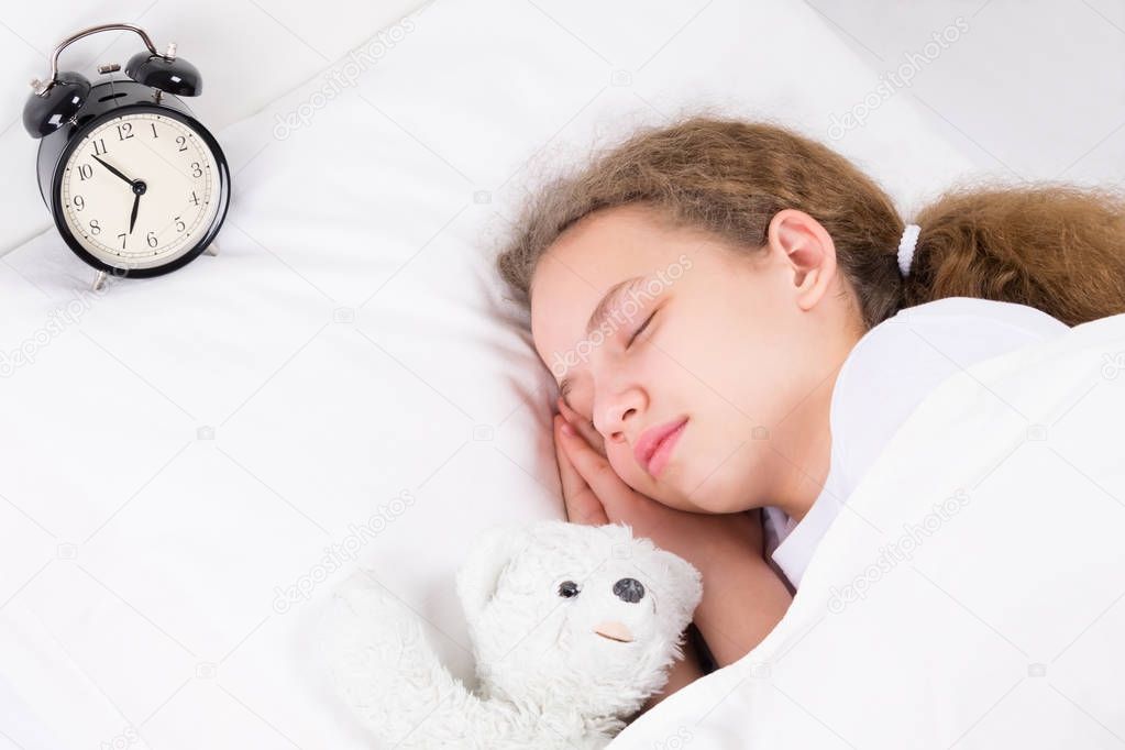 the girl is sleeping with an alarm clock, hugging a bear toy