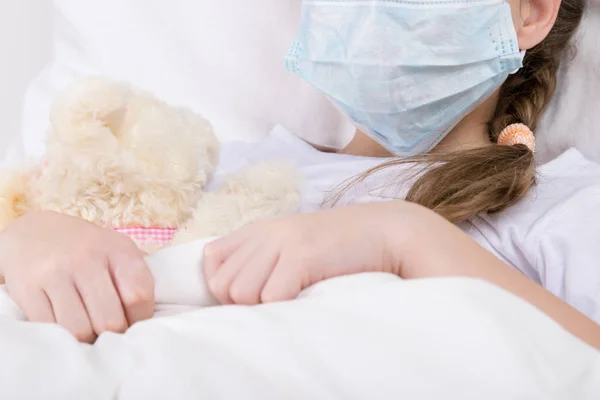 girl is sick with a mask on her face in bed and hugs a toy