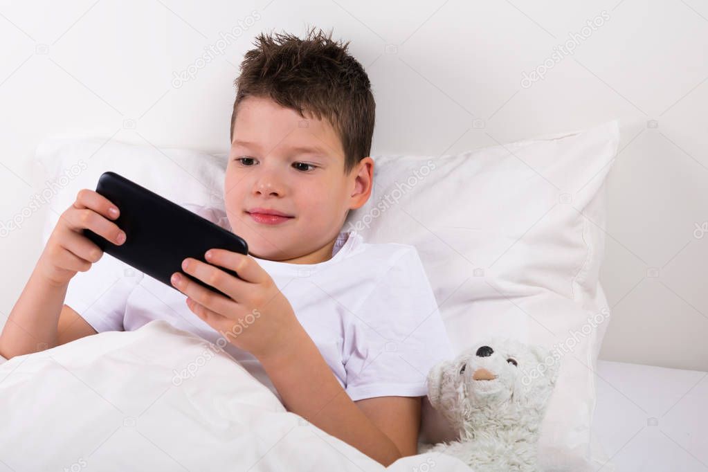 boy playing in the game on the phone lying in bed next toy white teddy bear
