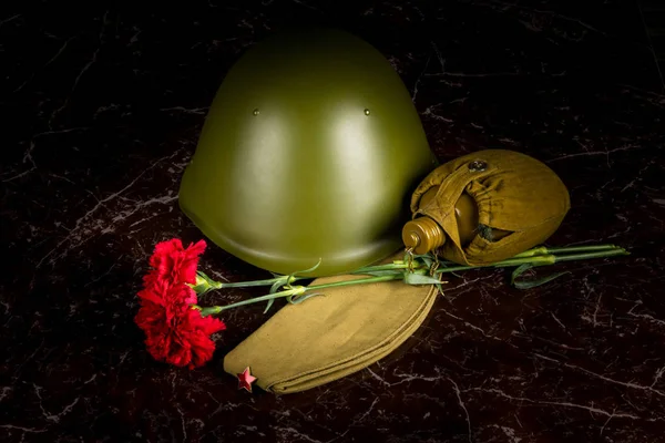 two carnations, a military helmet and a pilot\'s cap, a flask, against the background of the monument