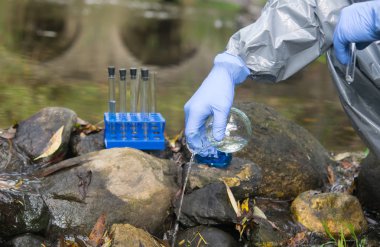 Bilim adamının eli koruyucu bir giysi ve eldivenle bir mataradan gölete sıvı döküyor.