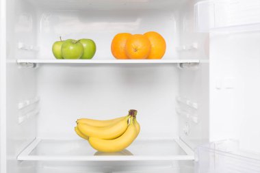 Taze yeşil elma, portakal ve olgun muzlar açık beyaz bir buzdolabında rafta yatıyor.