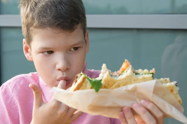 赤いシャツの少年が指を舐めてファーストフード店のおいしいサンドイッチを食べる — ストック写真