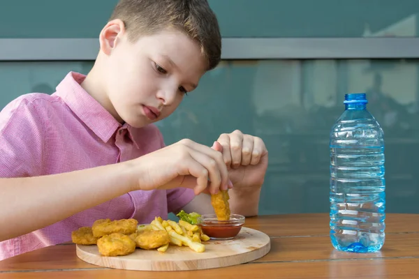 赤いシャツの少年がチキンナゲットをケチャップにつけて食べ ボトルから水を飲む — ストック写真