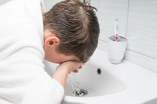 a boy in a white bathrobe in the bathroom washing his face with tap water above the sink