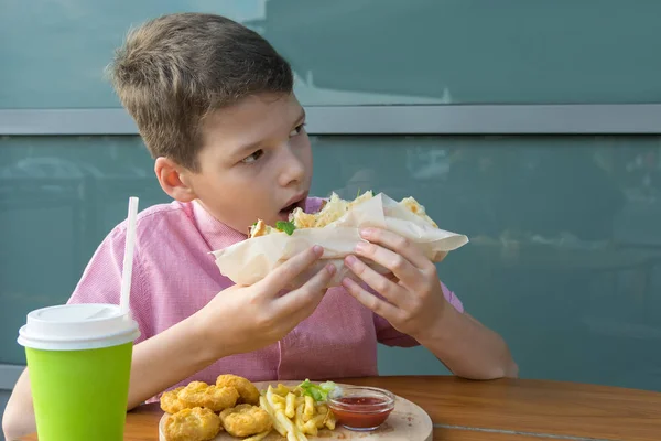 赤いシャツの男の子がおいしいサンドイッチを噛んでテーブルの上にはファーストフード店で買ったランチがあります — ストック写真