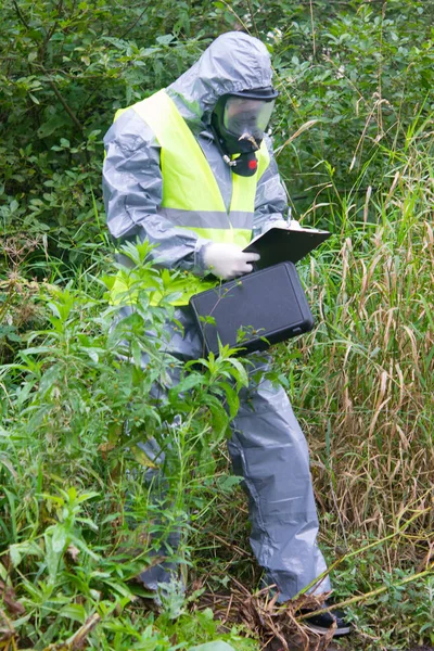 a specialist in a protective suit and mask, with a suitcase in his hands, records the results of the study, on a tablet, against the background of green nature